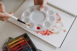 Close-up of hands holding a paintbrush and palette over an art notebook.
