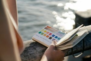 A person holding a watercolor journal with vibrant swatches by a shimmering waterfront.