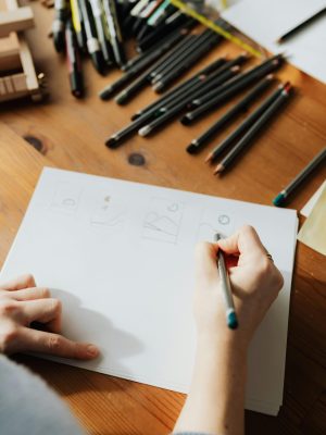pexels-photo-4219157-4219157 Close-up of a woman's hand sketching on paper surrounded by pencils on a wooden desk. Sketching pencil guide. How to use sketching pencils.