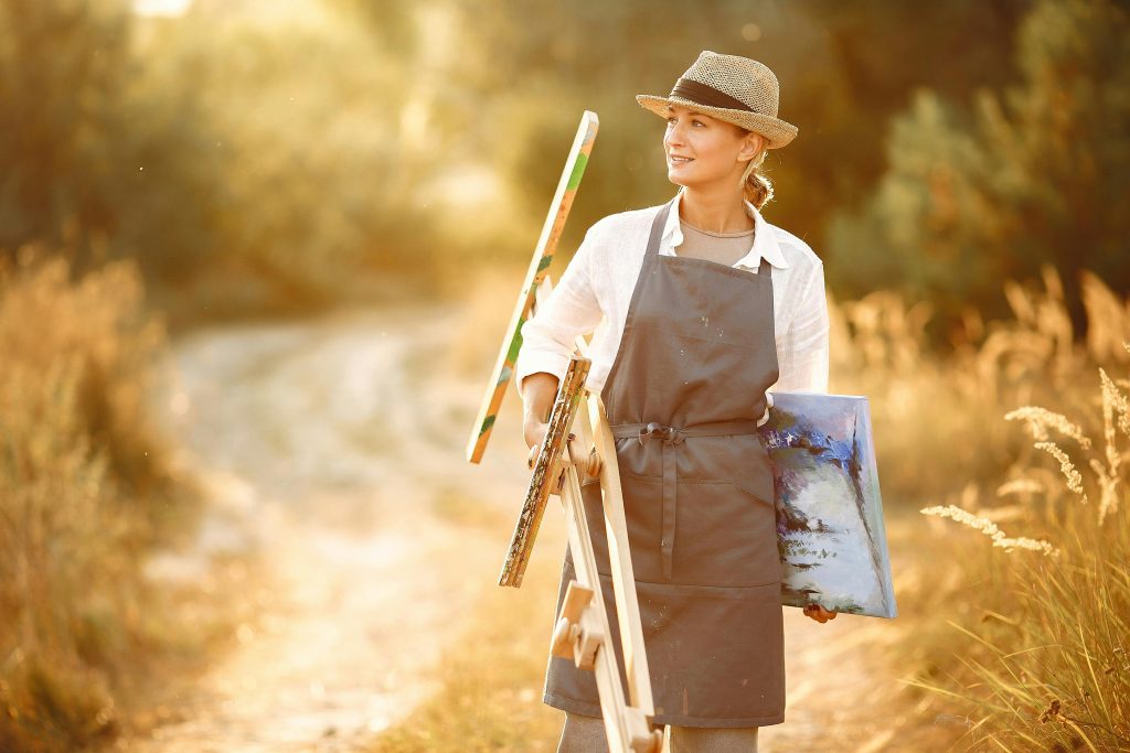 Happy female in hat and apron carrying easel and canvas while walking on road in summer sunny day in nature. painting techniques. painting styles.